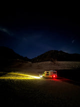 A bright-lit RV rental camper truck sits in a dark desert at night, illuminating sand. Mountains and faint stars dot the serene sky.