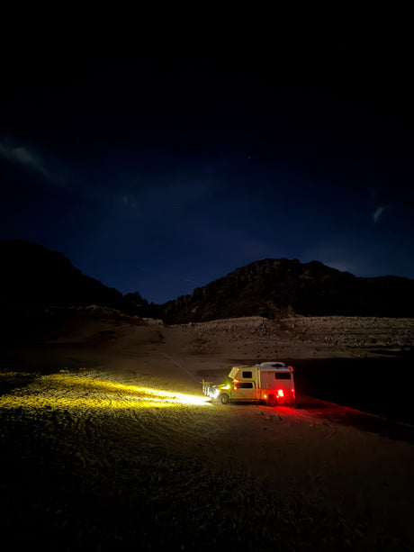 A bright-lit RV rental camper truck sits in a dark desert at night, illuminating sand. Mountains and faint stars dot the serene sky.