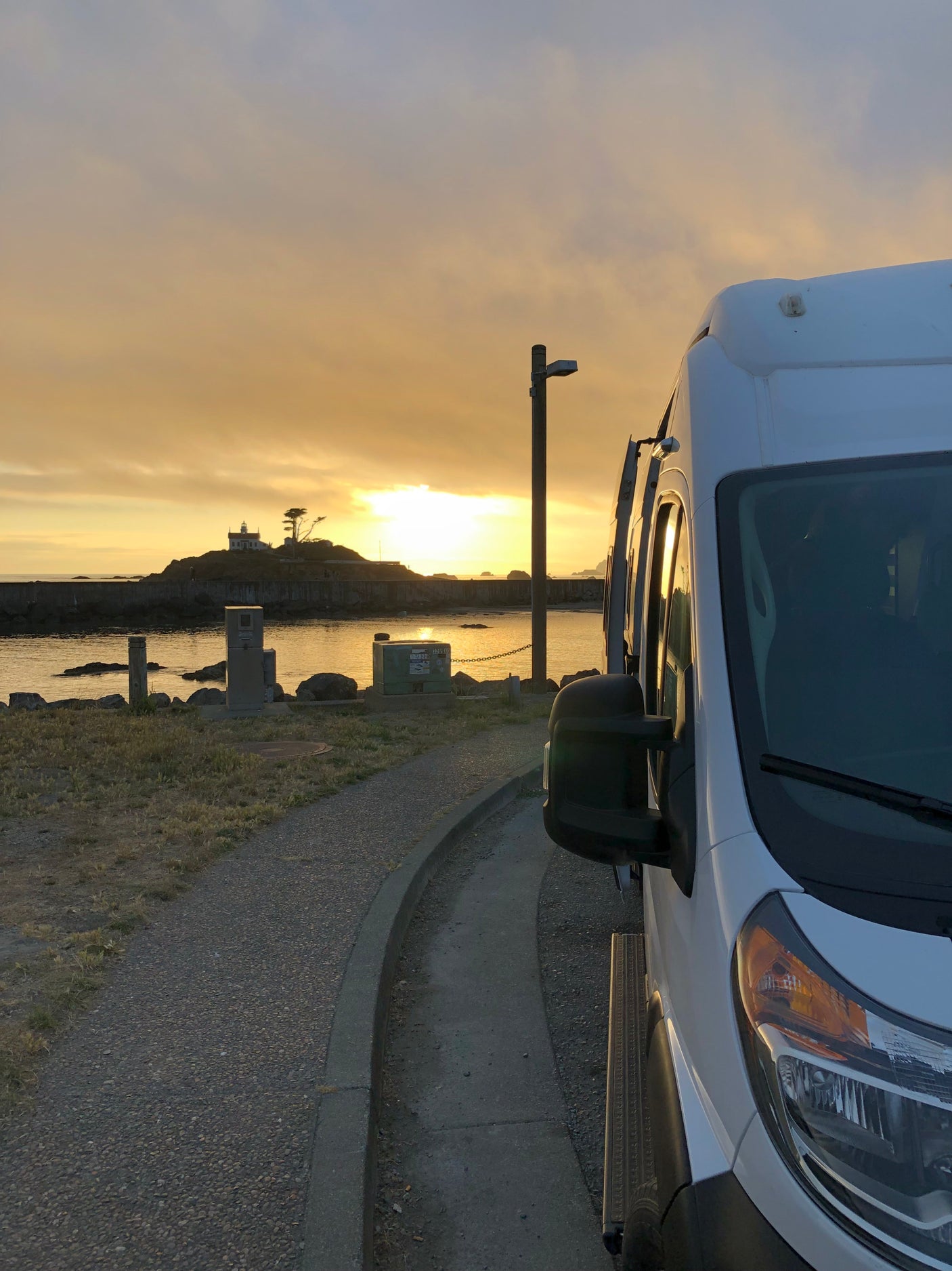 Enjoying a golden sunset over the ocean with a distant lighthouse from an RV rental, parked on a gravel path.