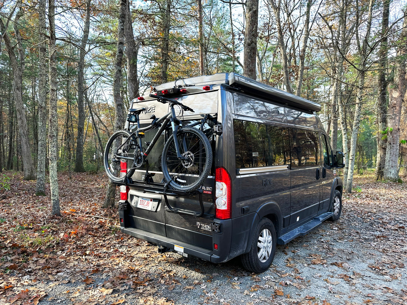 Dark grey RV rental, a camper van with two bikes on its rear rack, parked among trees with fallen autumn leaves.