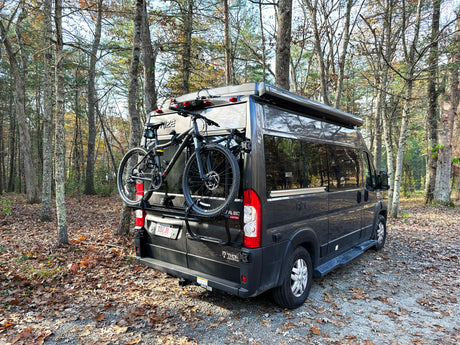 Dark grey RV rental, a camper van with two bikes on its rear rack, parked among trees with fallen autumn leaves.
