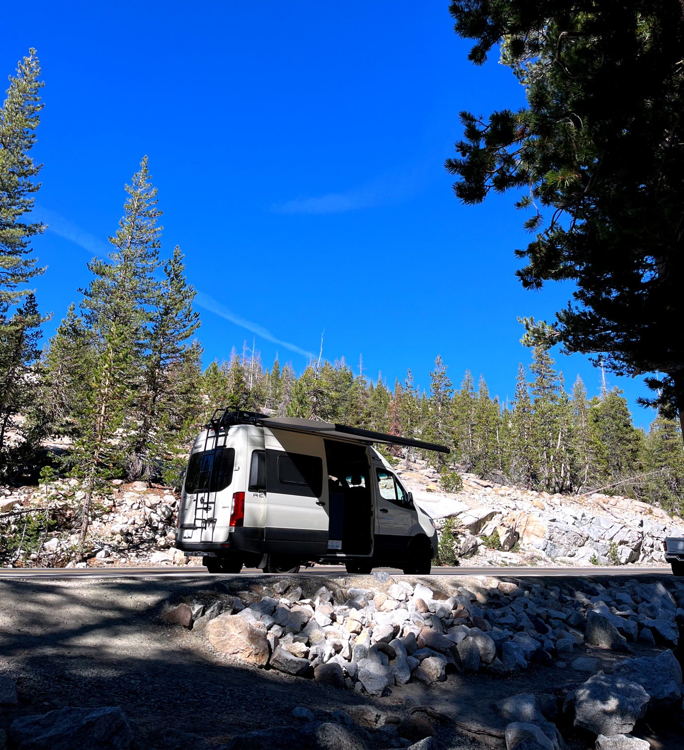 White RV rental parked on a sunny mountain road with extended awning. Pine trees & rocks surround this camper van adventure.