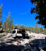 White RV rental parked on a sunny mountain road with extended awning. Pine trees & rocks surround this camper van adventure.