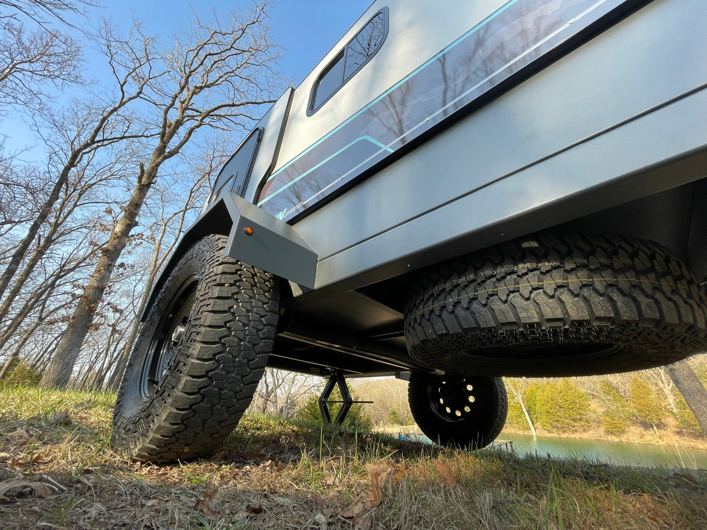 Low-angle shot of a rugged RV rental trailer with large tires and a spare, parked on grass by a serene lake.
