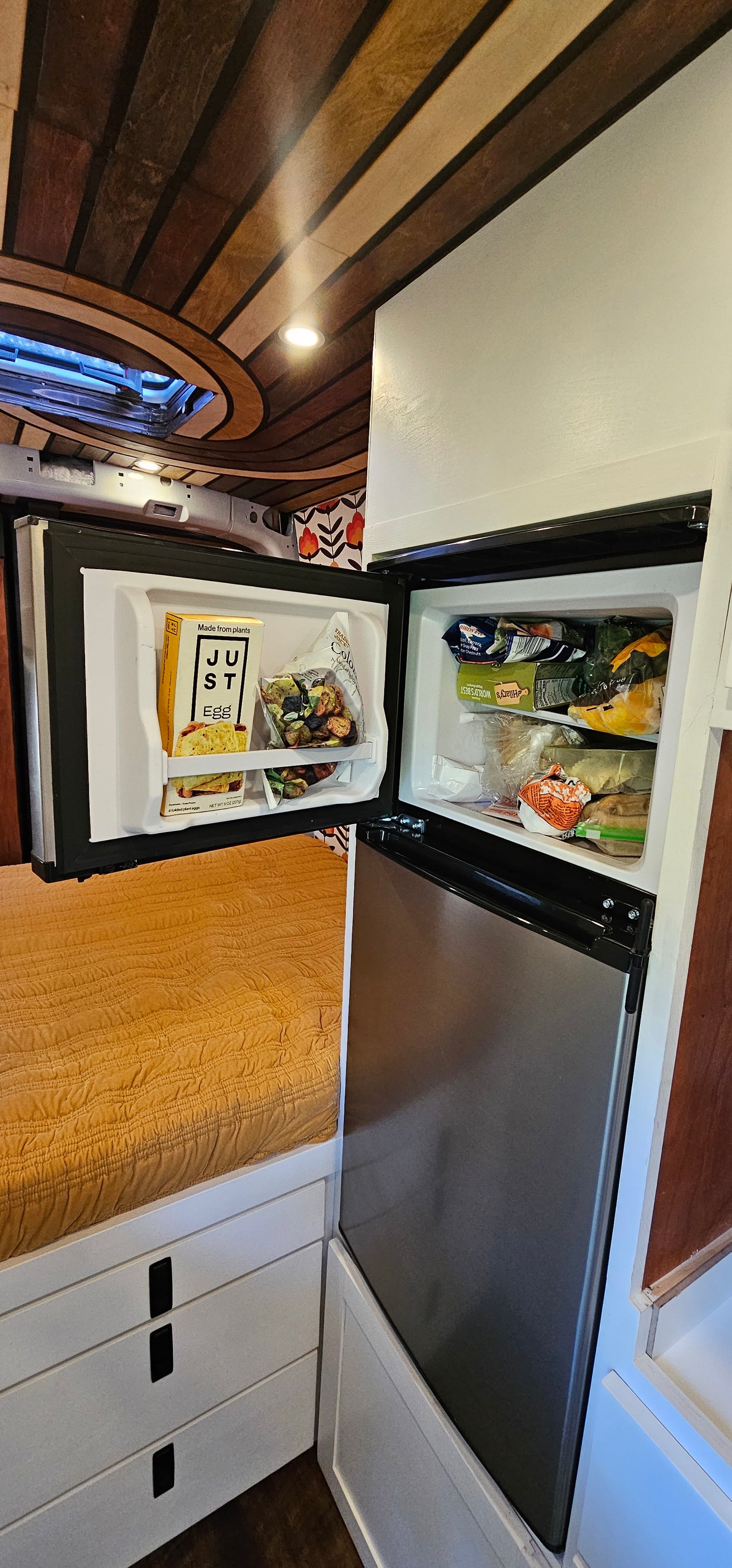 An RV rental's clever interior: Open fridge/freezer, storage drawers, & bed, all under a custom wood ceiling.