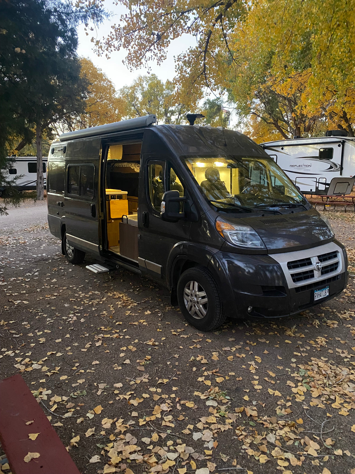 Dark gray camper van RV rental at an autumn campsite, side door open, warm interior visible, leaves scattered.