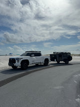 White Toyota Tundra pickup towing a dark gray CORE RV rental off-road trailer on an asphalt road by a sandy beach.