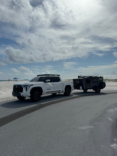 White Toyota Tundra pickup towing a dark gray CORE RV rental off-road trailer on an asphalt road by a sandy beach.