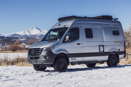 Silver Winnebago Revel 4x4 camper van rv rental parked on snow with mountains in background.