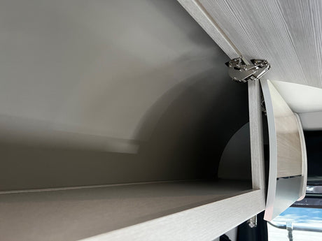 Open overhead storage cabinet in an RV rental, showing a curved door, light wood grain, and heavy-duty metal hinge.