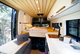 Modern rv rental interior: wood ceiling, comfy bed, kitchen, & laptop workspace with nature views.