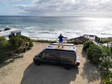 Coastal RV rental adventure: Man on top of a dark camper van, enjoying ocean views from his solar-powered setup.