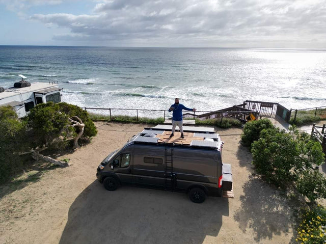  facing sunlit red mountains under a blue sky. Perfect for adventure.|Coastal RV rental adventure: Man on top of a dark camper van