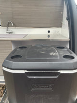RV rental compact kitchen area featuring a sink, countertop, and a black Coleman cooler for travel convenience.