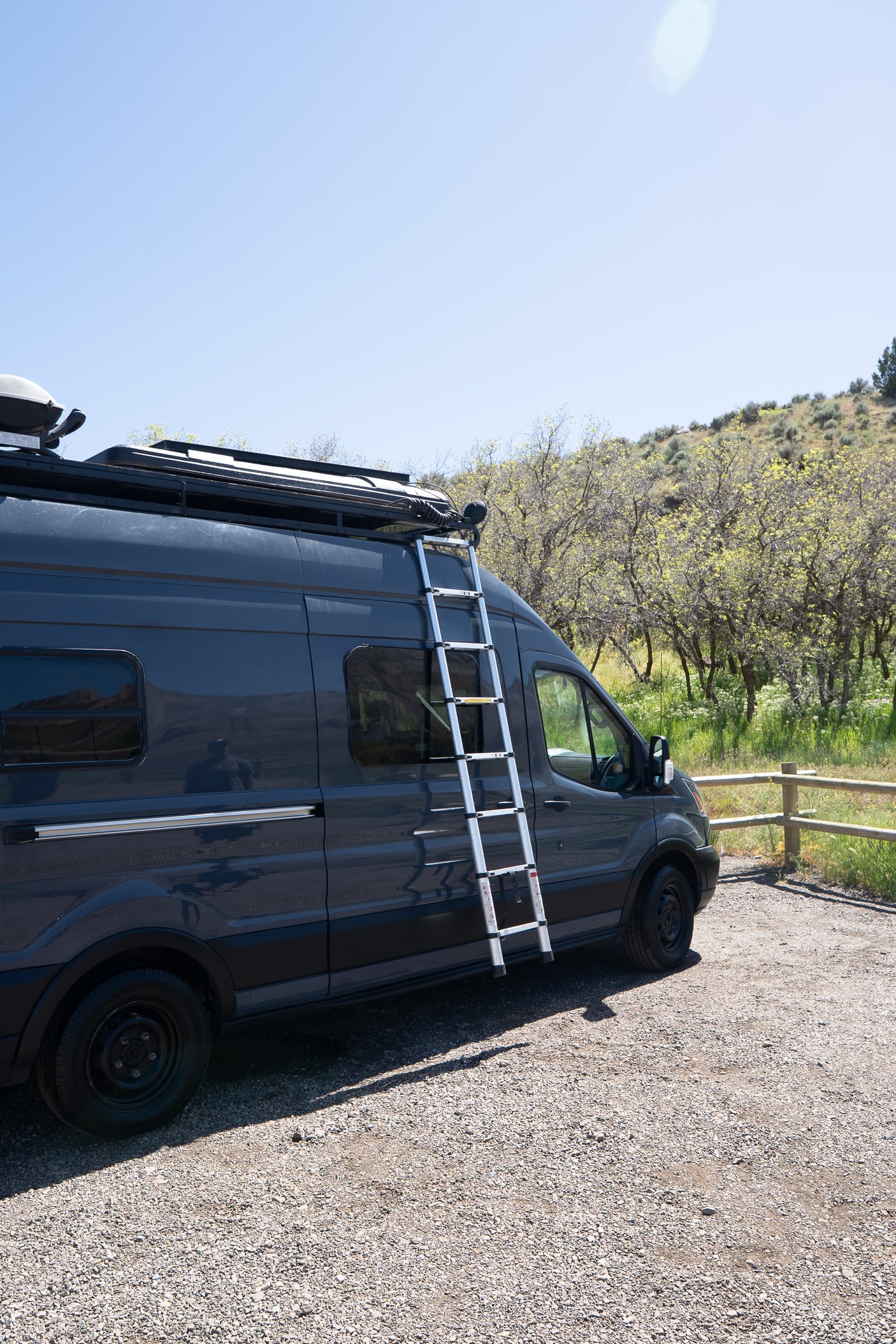 Dark grey rv rental campervan with a roof rack and ladder, parked on gravel next to trees under a clear blue sky.