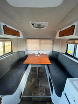 RV rental interior dinette features two gray benches, a wooden table, and windows with blinds, offering a cozy camper living space.