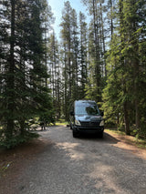 Dark RV rental van parked on a sunny gravel path in a serene pine forest, with a picnic table nearby.
