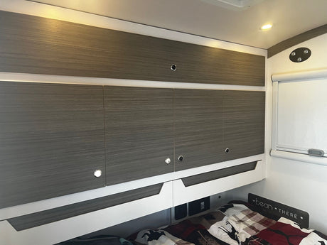 Modern RV rental interior: wood-grain overhead storage cabinets with integrated lighting, a bed, and bedside power ports.