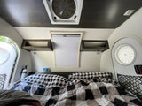 Inside a comfy RV rental: bed with plaid bedding, overhead shelves, & windows for natural light.