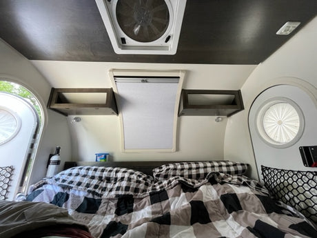 Inside a comfy RV rental: bed with plaid bedding, overhead shelves, & windows for natural light.