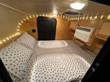 Cozy interior of an RV rental: a bed with turtle-patterned sheets, AC unit, & string lights. Ideal for travel.