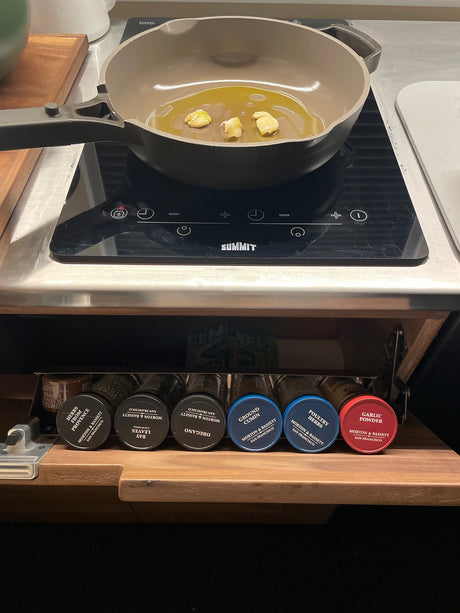 RV rental kitchen: Pan with garlic & oil on induction cooktop. Pull-out spice rack features Morton & Bassett herbs.