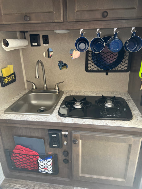 Compact kitchen in an RV rental, featuring a sink, two-burner stove, hanging mugs, and essential controls for water and power.