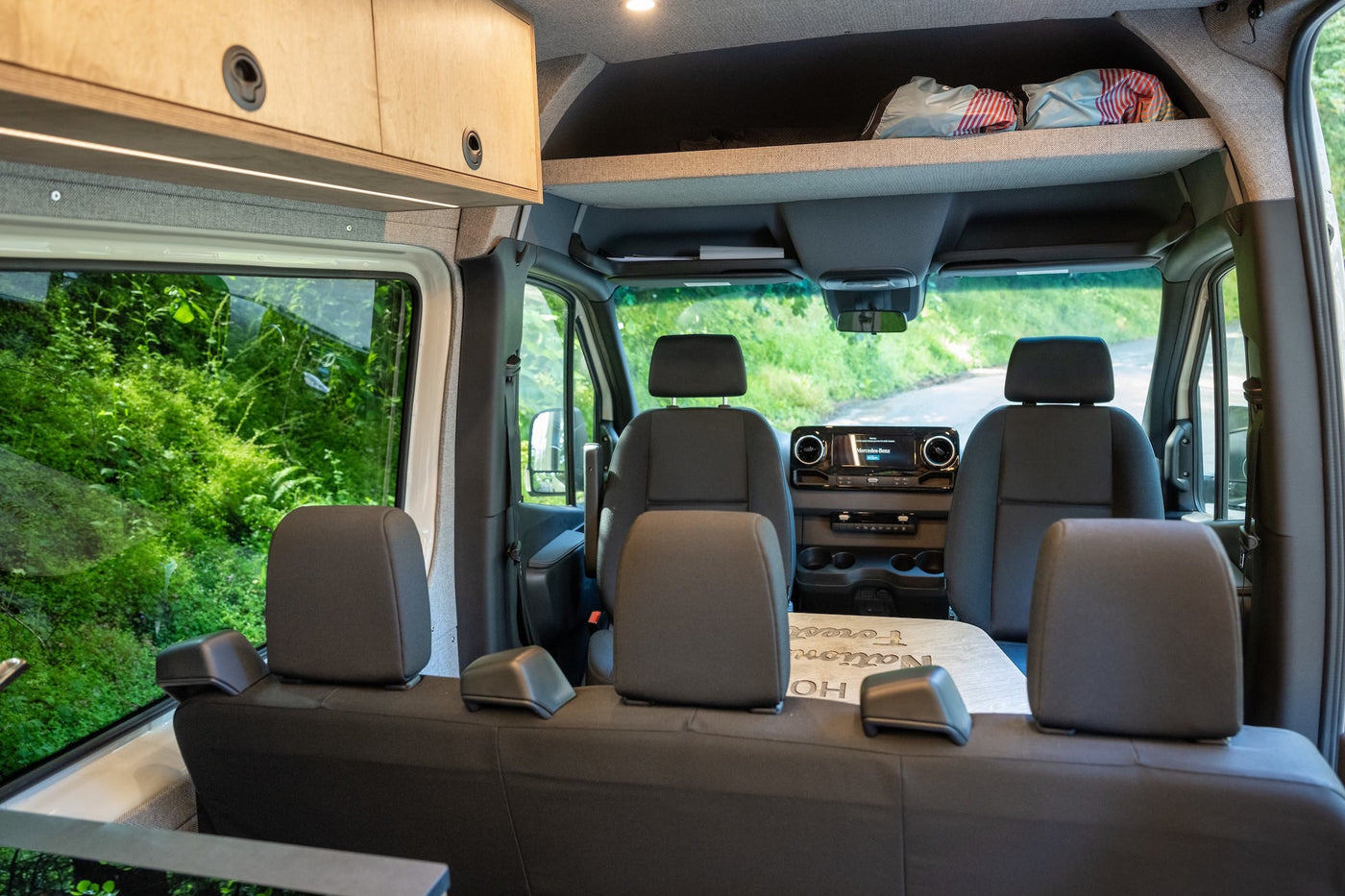 Spacious RV rental interior with custom cabinetry, overhead storage, front seats, and a lush green view outside. — photo 16
