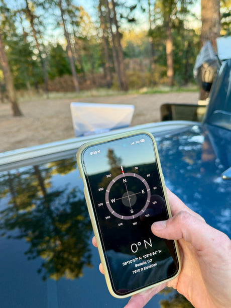 RV rental user holds a smartphone with a compass app showing 0°N, GPS coordinates for Sedalia, CO, 7910 ft elevation.