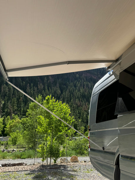 An RV rental with its awning extended creates shade at a sunny mountain campsite, surrounded by green trees and forest.