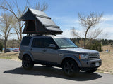 A gray Land Rover, fitted with a black rooftop tent, sits on asphalt by a scenic lake. Ideal for an RV rental outdoor adventure.