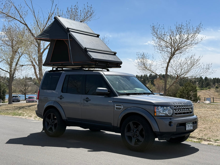 A gray Land Rover, fitted with a black rooftop tent, sits on asphalt by a scenic lake. Ideal for an RV rental outdoor adventure.