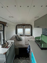 Interior of an RV rental showing a comfy bed, compact kitchen with sink/stove, and rear windows with a water view.