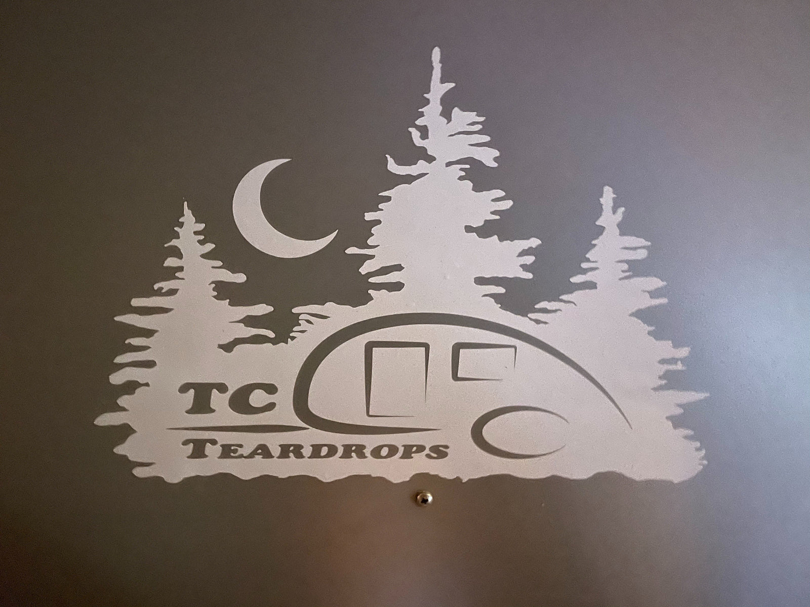 TC Teardrops RV rental graphic: A light silhouette of a teardrop trailer, pine trees, and moon on a dark wall.