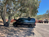 Dark RV rental camper van parked beneath large trees on a sunny day. Rural setting with stacked crates and farm trailers.
