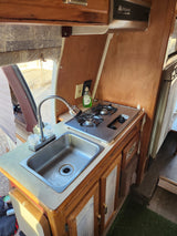 Inside an RV rental: a compact kitchen featuring a stainless steel sink, two-burner stove, and wooden cabinets.