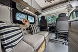 Bright RV rental interior shows lounge seating with striped pillows, flowers, and the front cabin view to nature.