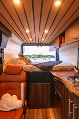 Inside this custom RV rental: wood-paneled ceiling, comfy bed, and a kitchen area, all with an ocean view.
