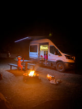 Family camping trip with an RV rental: Kids gather around a warm campfire beside their illuminated camper van at night.