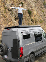 A happy woman on a gray customized RV rental van with rocky cliffs behind her, arms outstretched, celebrating travel.