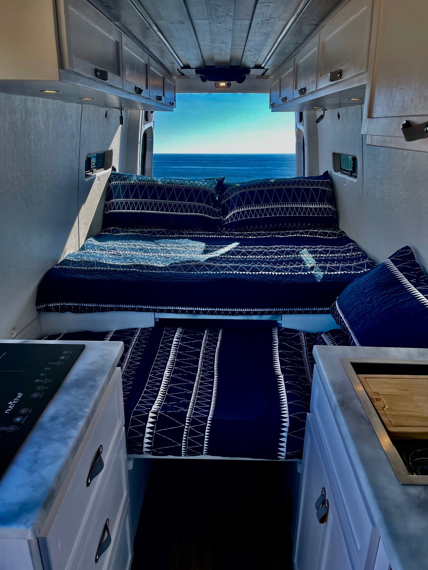 Step inside this RV rental! A comfy bed with blue bedding faces stunning ocean views, with a compact kitchen in the foreground. — photo 3