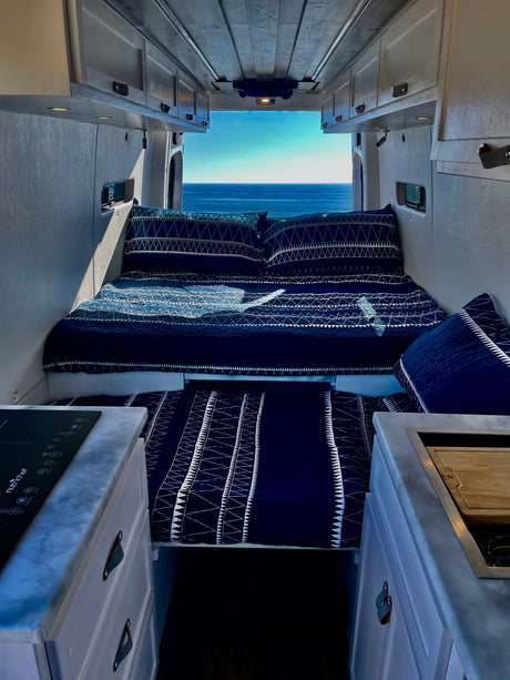 Step inside this RV rental! A comfy bed with blue bedding faces stunning ocean views, with a compact kitchen in the foreground.