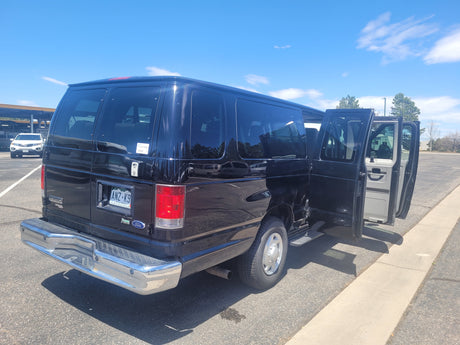 Rear of a black Ford van, doors open, parked on asphalt near RV rental storage on a sunny day.