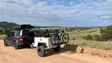 Overlanding RV rental setup: SUV with roof tent, trailer & bike on a dirt road amidst scenic mountains. Ready for adventure!