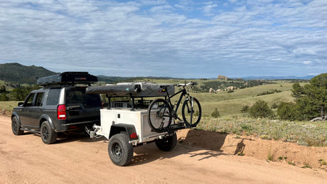 Overlanding RV rental setup: SUV with roof tent, trailer & bike on a dirt road amidst scenic mountains. Ready for adventure!