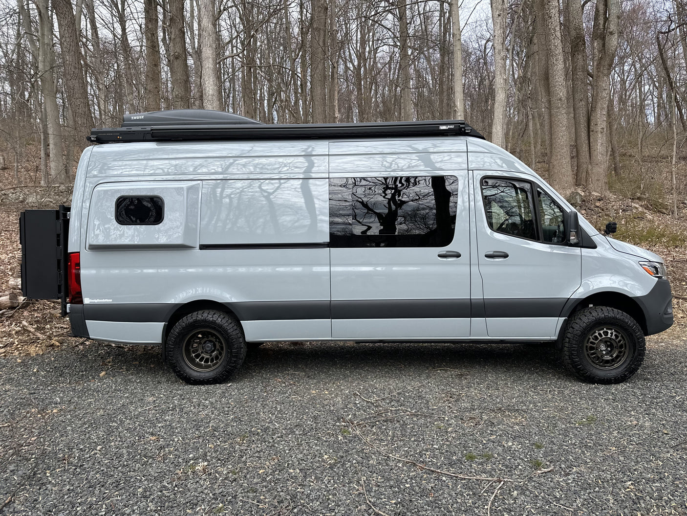 This light grey Sprinter camper van, ideal for RV rental, is parked on gravel with off-road tires and roof storage.