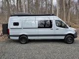 This light grey Sprinter camper van, ideal for RV rental, is parked on gravel with off-road tires and roof storage.
