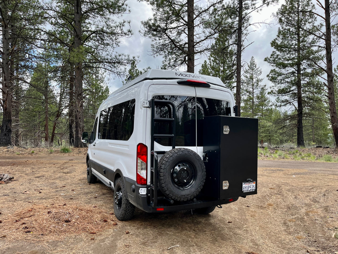White ModVans **rv rental** camper van parked in a pine forest. Rear view with spare tire, ladder, and storage box.