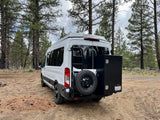 White ModVans **rv rental** camper van parked in a pine forest. Rear view with spare tire, ladder, and storage box.