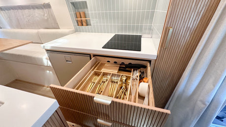 Inside an RV rental: a modern kitchenette with light blue tiled backsplash, built-in fridge, and organized golden flatware in a pull-out drawer.